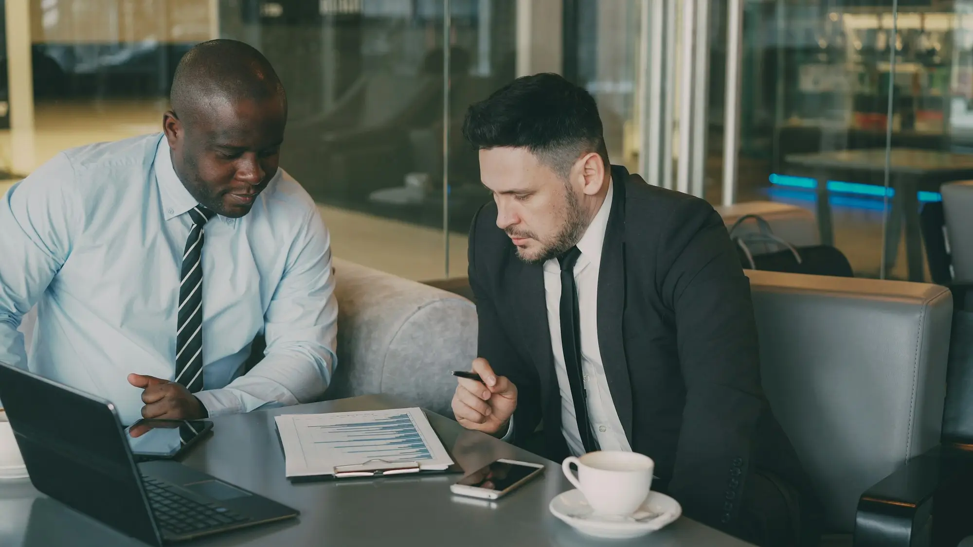 Two business owners at a café table reading through a printed loan contract beside a laptop, one holding a pen